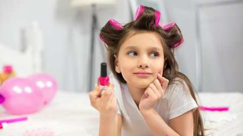 Preschool-aged-girl-lying-on-a-white-blanket-with-hair-curler-holding-nail-polish