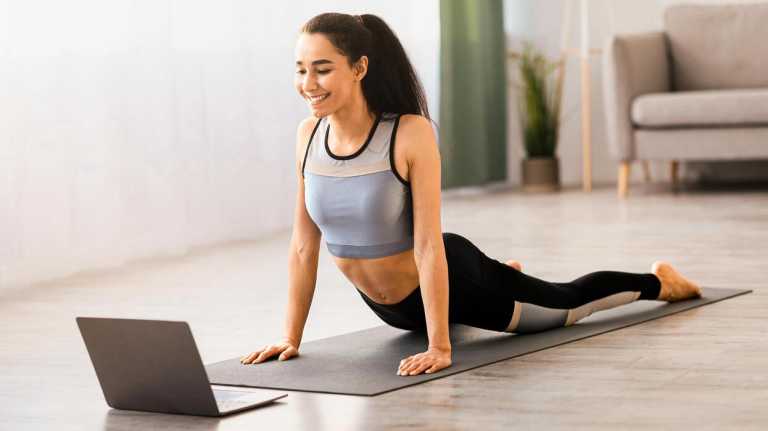 fitness-online-athletic-young-woman-doing-yoga-in-living-room-stretching-on-yoga-mat-in-front-of-laptop-copy-space-good-looking-brunette-lady-exercising-at-home-using-lessons-side-view-photo